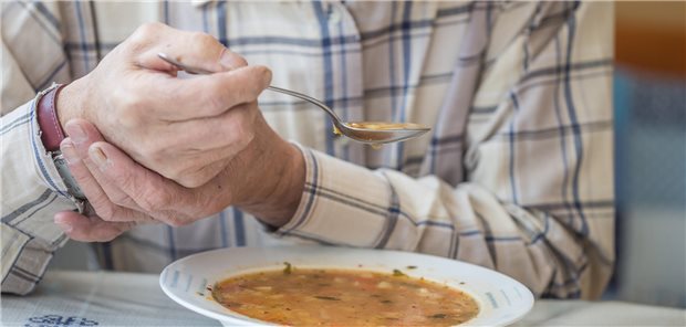 Älterer Mann isst Suppe und hält seine Hand mit der anderen Hand fest.