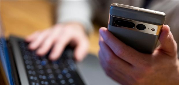 Ein Mann sitzt am Laptop und hält ein Smartphone mit in der Hand.