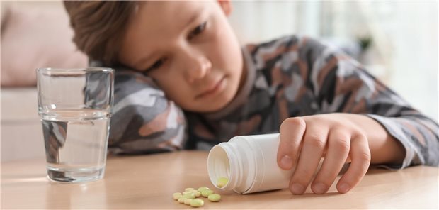 Ein kleienr Junge mit Tabletten und Wasser an einem Tisch im Haus, repräsentiert die Gefahr einer Medikamentenvergiftung.