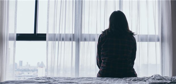 Eine Frau sitzt auf einem Bett und schaut gedankenverloren aus dem Fenster.