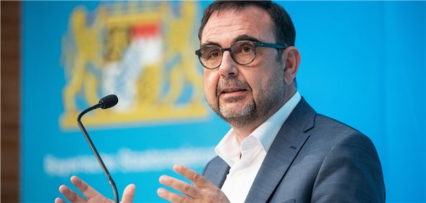 Mit Leasinghühnern Demenzkranken helfen Bayerns Gesundheitsminister Klaus Holetschek vor dem undeutlichen großen Wappen Bayerns am Mikrofon mit einem freundlichen Gesichtsausdruck und gehobenen Händen.
