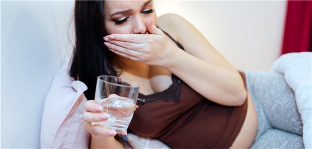 Schwangere, die auf der Seite liegt, sich mit einem unglücklichen Gesichtsausdruck die Hand vor den Mund hält, während sie nach einem Glas Wasser greift.