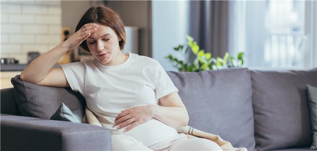 Migräne in der Schwangerschaft: Welche Therapie ist überhaupt möglich? Eine schwangere Frau sitzt auf dem Sofa und hält sich eine Hand auf die Stirn.