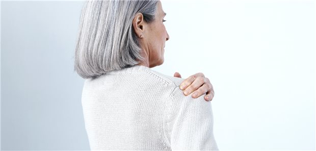 Eine ältere Frau mit langen grauen Haaren greift sich an die Schulter. 