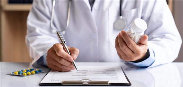 Ein männlicher Arzt hält eine Flasche mit Tabletten in der Hand und füllt an seinem Arbeitstisch ein Rezept für den Patienten aus.