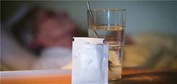 Eine Tüte vor einem Glas Wasser, im Hintergrund liegt eine alte Frau im Bett.