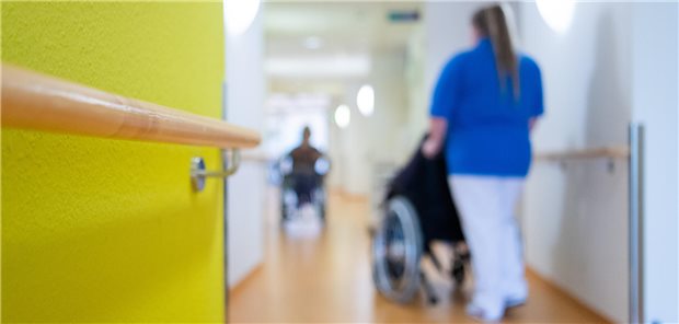 Frau schiebt einen Mann im Rollstuhl über einen Krankenhausflur.