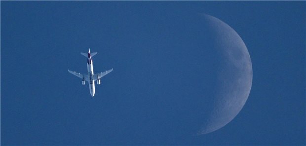 Ein Flugzeug wird von unten im Flug fotografiert. Neben dem Flieger ist der sichelförmige Mond.