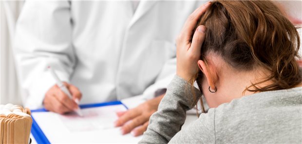 Eine Patientin schlägt die Hände auf dem Kopf zusammen. Gegenüber sitzt ein Arzt in weißem Mantel.