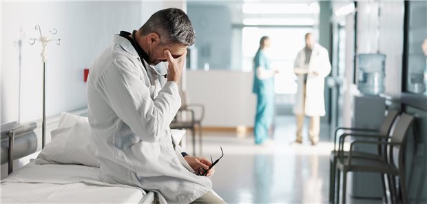 Ein Klinikarzt sitzt erschöpft auf einem Bett im Flur und hält sich mit einer Hand den Kopf.