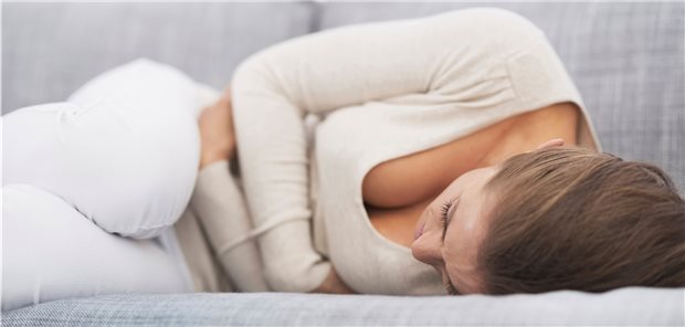 Frau liegt, sich vor Schmerzen krümmend, auf einem Sofa, die Arme auf den Bauch gepresst.