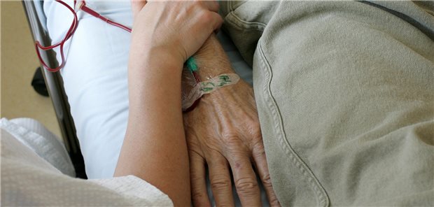 Eine junge Person hält die regungslose hand einer älteren Person, die in einem Krankenhausbett liegt.