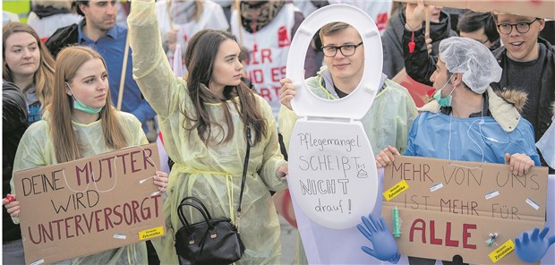 Pflegekräfte im Öffentlichen Dienst legen Arbeit nieder „Deine Mutter wird unterversorgt“ steht auf einem Schild bei einem Warnstreik in der Münchner Innenstadt. Nebendran ein Mann, der eine Klobrille mit der Aufschrift „Pflegemangel – Scheißt nicht drauf!“ um den Hals trägt.