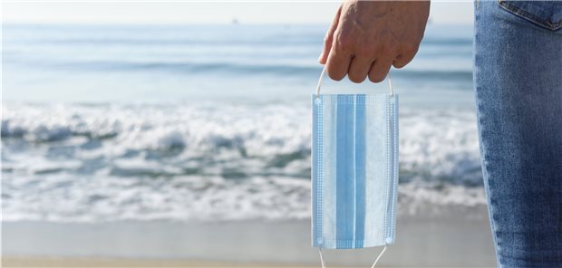 Corona: Sommer, Sonne, Delta-Variante? Person am Meer, in der Hand eine Schutzmaske.