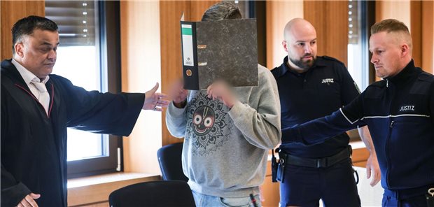Der angeklagte Krankenpfleger hält sich zwischen Anwälten und Justizangestellten einen Aktenordner vor sein Gesicht, während er zu seinem Platz im Aachener Landgericht gebracht wird.