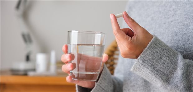 Nahaufnahme einer Frau mit einer weißen Pille in der einen und einem Glas Wasser in der anderen Hand. 