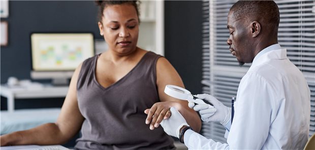Seitenansicht eines afroamerikanischen Arztes, der im Rahmen einer Untersuchung die Haut einer Patientin in einer Klinik durch ein Vergrößerungsglas betrachtet.