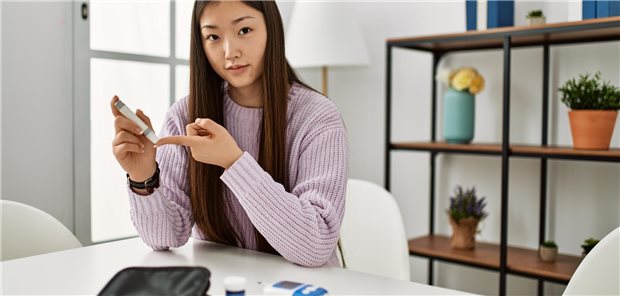 Junges Mädchen mit asiatischem Migrationshintergrund sitzt am Tisch und misst ihren Blutzucker.