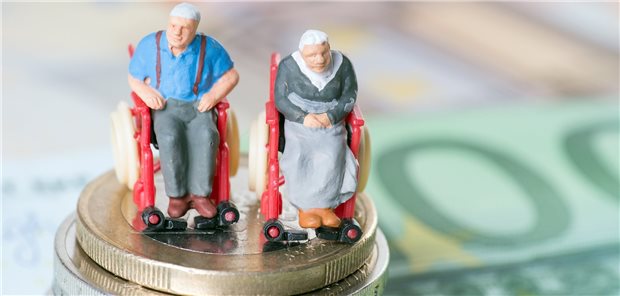 Sozialhilfeausgaben bei Pflege gehen steil nach oben Figuren eines älteren Herren und einer älteren Frau in Rollstühlen auf einem Stapel Euro-Münzen, im Hintergrund unscharf verschiedene Euro-Scheine.