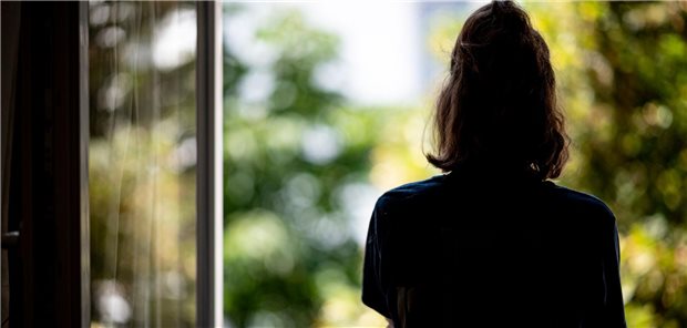 Eine Frau steht am Fenster und guckt raus. Sie ist von hinten zu sehen.