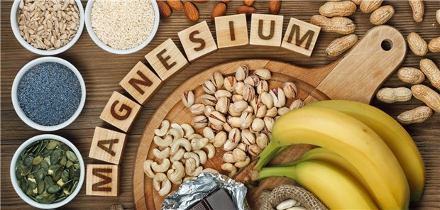 Magnesiumhaltige Lebensmittel wie Bananen, Mohn, Cashew-Kernen und Erdnüsse liegen auf dem Tisch, dazwischen das Wort &quot;Magnesium&quot; auf Holzklötzchen.