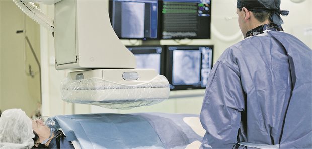 Das Bild zeigt einen Blick in ein Herzkatheterlabor. Ein Patient wird mit bildgebenden Instrumenten untersucht. Ein Arzt betrachtet die Ergebnisse auf dem Bildschirm.
