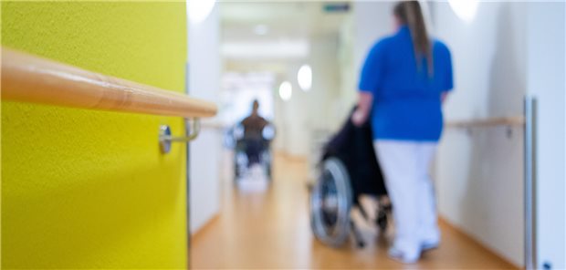 Eine Pflegerin schiebt eine Bewohnerin eines Pflegeheims in einem Rollstuhl über den Flur.