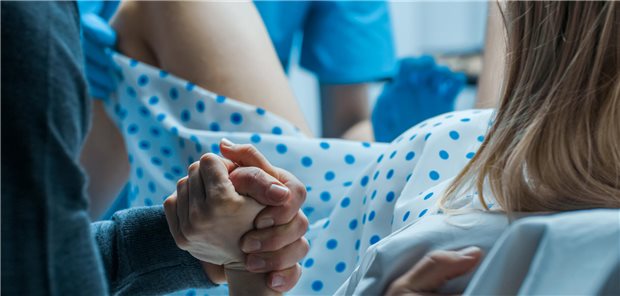 Nahaufnahme einer Frau bei der Geburt, während ihr Ehemann ihr zur Unterstützung die Hand hält und ein Geburtshelfer bei der Entbindung assistiert.