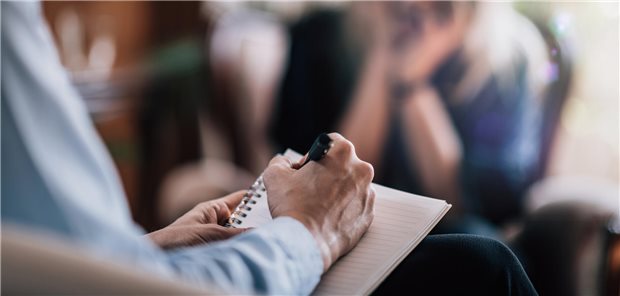 Ein Psychoterapeut spricht mit einer Patientin un dhält einen Block und Stift in der Hand.