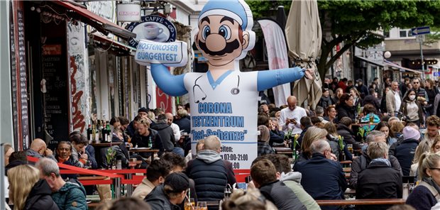 Kostenfreie Corona-Tests im Sommer – Kommunen dafür, Marburger Bund dagegen Die Straßencafes im Hamburger Schanzenviertel sind mit Besuchern gefüllt, während eine Hinweisfigur für ein ebenfalls dort beheimatetes Corona-Testzentrum auf dessen Eingang hinweist. Die letzten Corona-Maßnahmen sind in Hamburg weggefallen.