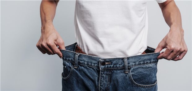Ein junger Mann zeigt wieviel Gewicht er verloren hat, indem er sich in seine alten übergroßen Jeans stellt und somit zeigt wie sehr sich sein Bauchumfang reduziert hat.
