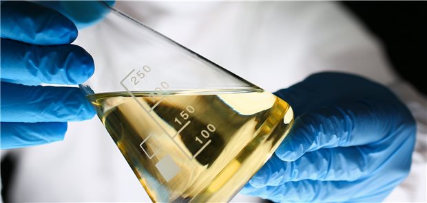 Erlenmeyer-Kolben mit gelblicher Flüssigkeit hält eine Person mit blauen Gummihandschuhen in einem weißen Kittel schräg (Symbolbild).