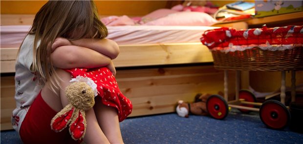 Ein Kind sitzt am Boden seines Kinderzimmers und hat die arme vor dem Gesicht verschränkt als würde es weinen.