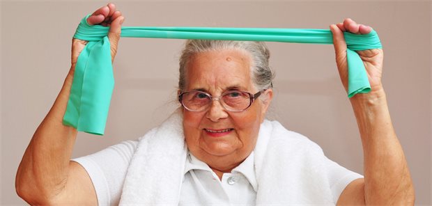 Das Bild zeigt eine ältere Dame, die eine leichte Übung macht. Sie spannt ein Tuch mit den Händen über ihrem Kopf.