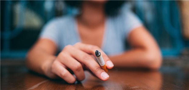 Frau hält eine Zigarette in die Kamera und sitzt an einem Tisch