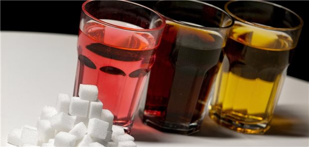 Drei Gläser mit verschiedenen Limonaden stehen auf einem Tisch, davor liegen mehrere Würfel Zucker. 