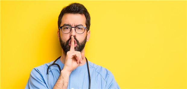 Ein Arzt hält sich den Zeigefinger vor den Mund um Ruhe anzudeuten. 