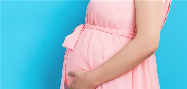 Schwangere Frau im rosa Kleid hält sich den Bauch und steht vor einem blauen HIntergrund.