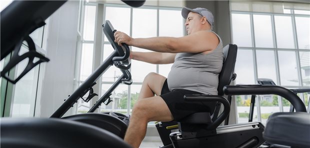 Ein übergewichtiger Mann sitzt auf einem Standfahrrad im Fitnessstudio. 