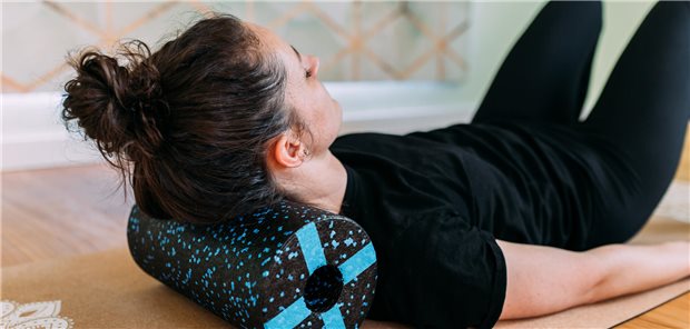 Frau in schwarzer Sportkleidung liegt mit einer Faszienrolle unter dem Nacken auf einer Yogamatte und macht Übungen zur Stärkung der Nackenmuskulatur.