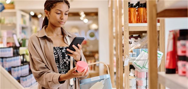 Eine junge Frau überprüft beim Einkauf in einer Kosmetikboutique mit ihrem Smartphone die Inhaltsstoffe einer Creme.