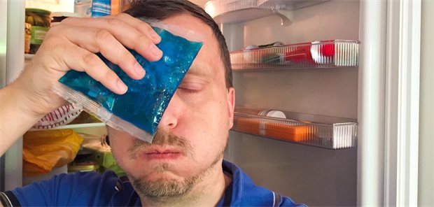 Mann kühlt sich mit Kühlpad die Stirn vor offenem Kühlschrank. 