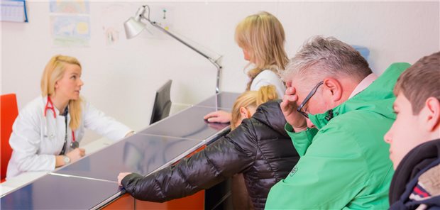 Hohe Arbeitsbelastung der MFA: Welche Strategien sich lohnen Eine Ärztin sitzt hinter dem Anmeldepult. Es warten viele Patientinnnen und Patienten in einer Schlange.