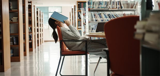 Eine Frau sitzt am Tisch und legt sich ein Buch aufs Gesicht.