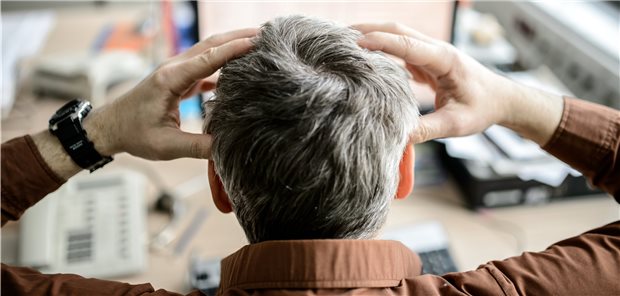 Überarbeitung führt nicht direkt zu Berufskrankheit Person greift sich an den kopf - vor ihr ein Bildschirm und Tastatur, ein Telefon sowie ein mit Dokumenten beladener Ordner.