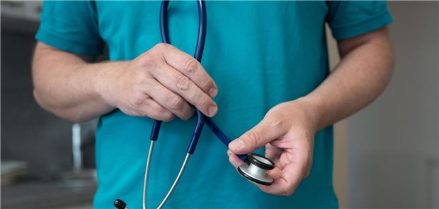 Ein Hausarzt steht in seiner Hausarztpraxis und hält ein Stethoskop. 