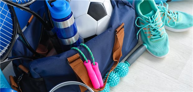 Eine Sporttasche liegt am Boden und ist gefüllt mit Sportsachen, wie einem Springseil, einem Fußball, Laufschuhen und Tennisschlägern. 