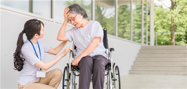 Eine ältere Frau im Rollstuhl fasst sich mit der Hand an den Kopf. Eine Pflegerin sitzt daneben und kümmert sich. 