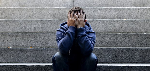 Ein junger Mann sitzt auf einer Stufe und hält sich die Hände vor das Gesicht.