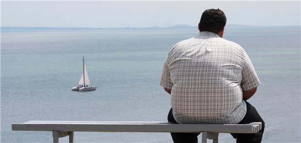 Korpulenter Mann auf einer Bank vor dem Meer.